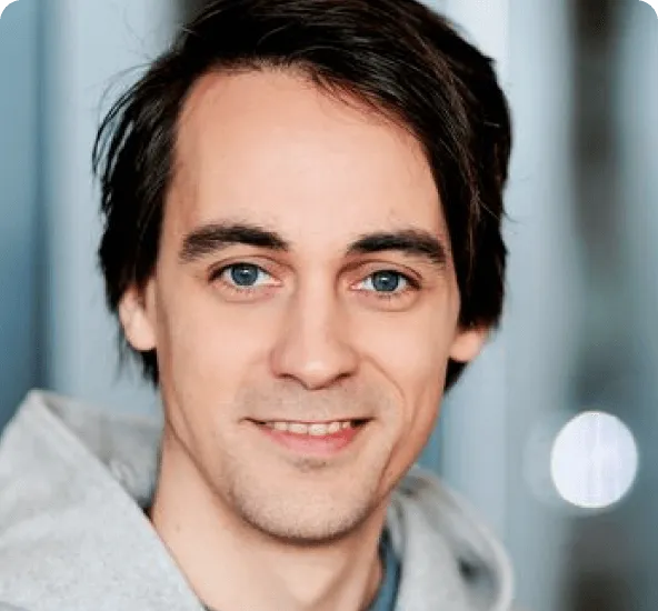 Close-up of a smiling man with dark hair and blue eyes wearing a light gray hoodie.